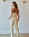 A woman stands with her back to the camera wearing a light beige strapless tube top and matching wide-leg trousers, paired with metallic heels, in a minimalist room with a white wall and gold wall art.