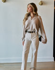 A woman stands in a room wearing a shimmering champagne long-sleeve knit sweater, off-white wide-leg trousers, a black belt, and black pointed shoes. She is holding a small black handbag and wearing layered gold necklaces.