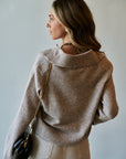 A woman wearing a light taupe knit sweater and cream pants, shown from a side-back angle against a plain light gray wall. She is carrying a small black shoulder bag with a gold chain strap.