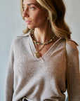 A woman wearing a light beige knit V-neck sweater, layered gold necklaces, and a black belt, shown in a close-up lifestyle shot against a neutral light gray background.