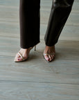 A close-up of a person's feet wearing metallic gold strappy high-heeled sandals and dark brown pants, standing on a light gray wood plank floor.