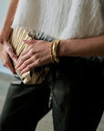 A close-up lifestyle shot of a person holding a gold pleated clutch bag, wearing a white fuzzy top, black pants, and gold bangles.