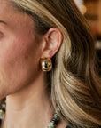 A close-up of a woman's ear wearing a chunky, polished gold dome stud earring, with her blonde hair and a beaded necklace visible.