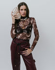 Woman wearing a patterned sheer top and maroon pants holding a shell clutch against a plain background