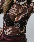 model wearing a patterned sheer top with a necklace and maroon pants on a plain background