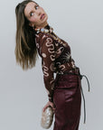 Woman wearing a patterned sheer top and maroon pants against a plain background side view
