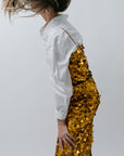 Person wearing a white shirt and gold sequin dress against a plain background side view