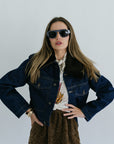 Woman wearing sunglasses, a denim jacket, and a patterned shirt against a white background