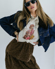 Woman wearing a denim jacket, white graphic t-shirt, and brown skirt with sunglasses on a plain background