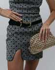Person wearing a black and white patterned dress with a belt, holding a gold clutch against a gray background