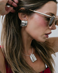 Woman wearing sunglasses, Large Sundra Pendant Necklace Silver and silver earrings with a neutral background