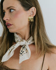 Woman wearing a gold earring and a white scarf with black patterns.