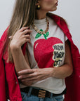 Woman wearing a white t-shirt with a red apple graphic and 'New York City' text, paired with a red jacket, close up front view