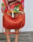 Red woven handbag held by a person wearing a colorful floral dress on a wooden floor.
