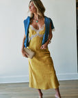 A woman models a yellow midi slip dress with a lace-trimmed neckline, styled with a blue shirt over her shoulders, a gold clutch, and gold high-heeled sandals against a white wall.