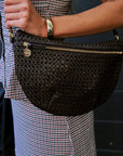 A close-up of a person wearing a dark brown woven faux-leather half-moon shoulder bag with gold-tone hardware, paired with a red, white, and black gingham set.