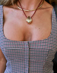A close-up of a person wearing a gold shell-shaped pendant necklace on a burgundy cord, layered with Cleopatra Chain, over a black, white, and red gingham top.