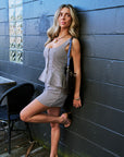 A woman posing against a dark gray wooden wall, wearing a sleeveless, mini-length checkered set with a peplum waist and a small shoulder bag.