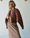 A woman models a brown cropped bomber jacket over a brown and beige patterned top and matching skirt, standing against a white wall with decorative gold wall hangings.