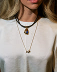 A close-up of a woman wearing layered necklaces, including a black cord choker with a gold teardrop pendant containing blue and white stones, and a delicate gold chain necklace with small gold charms.