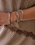 A close-up of a person's wrist wearing three silver-toned bangles, including a textured flat band, a small bead-joined loop, and a larger open-ended bangle, set against a brown and beige gingham patterned garment.