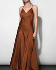 A woman modeling a floor-length satin slip dress in a warm rust brown color, standing against a plain light gray wall.