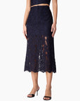 A model wearing a dark navy blue lace midi skirt with a scalloped hem, paired with a matching crop top and black heeled sandals, shown against a plain white background.