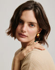 Woman with short brown hair wearing wood and gold earrings and a beige sweater against a light background