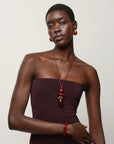 A model wearing a dark plum strapless dress, a red beaded necklace, a red bangle, and red hoop earrings, posed against a light gray studio background.