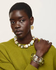 A model with short dark hair is shown from the chest up, wearing a textured olive green knit sweater, a chunky beaded necklace with white, cream, and light green beads, gold-toned earrings, and a clear acrylic bangle. The background is a clean, solid off-white studio backdrop.