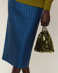 A close-up of a person wearing a blue quilted midi skirt and holding a small metallic gold fringe tassel bag against a plain light beige background.