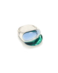 A modern sculptural ring featuring a wide, polished silver band with a large, organic-shaped inlay of translucent blue and green glass, set against a clean white background.