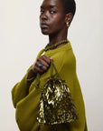 A model wearing a textured olive green sweater and matching green braided accessories, holding a gold sequined fringe handbag against a plain light background.
