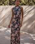 A woman wearing a sleeveless, high-neck, floor-length black dress with a pink and beige floral paisley pattern, standing outdoors against a beige stone wall with dappled sunlight.