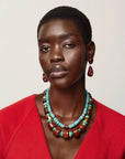 A close-up portrait of a model wearing a multi-strand turquoise and brown beaded necklace with matching drop earrings, paired with a bright red top against a light beige background.