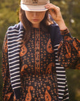 A woman outdoors wearing a beige baseball cap, a navy and orange paisley set, and a navy striped cardigan.
