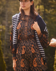 A woman wearing a dark paisley set with orange motifs, a navy and white striped cardigan, and a beige baseball cap, standing outdoors in a blurred forest setting.