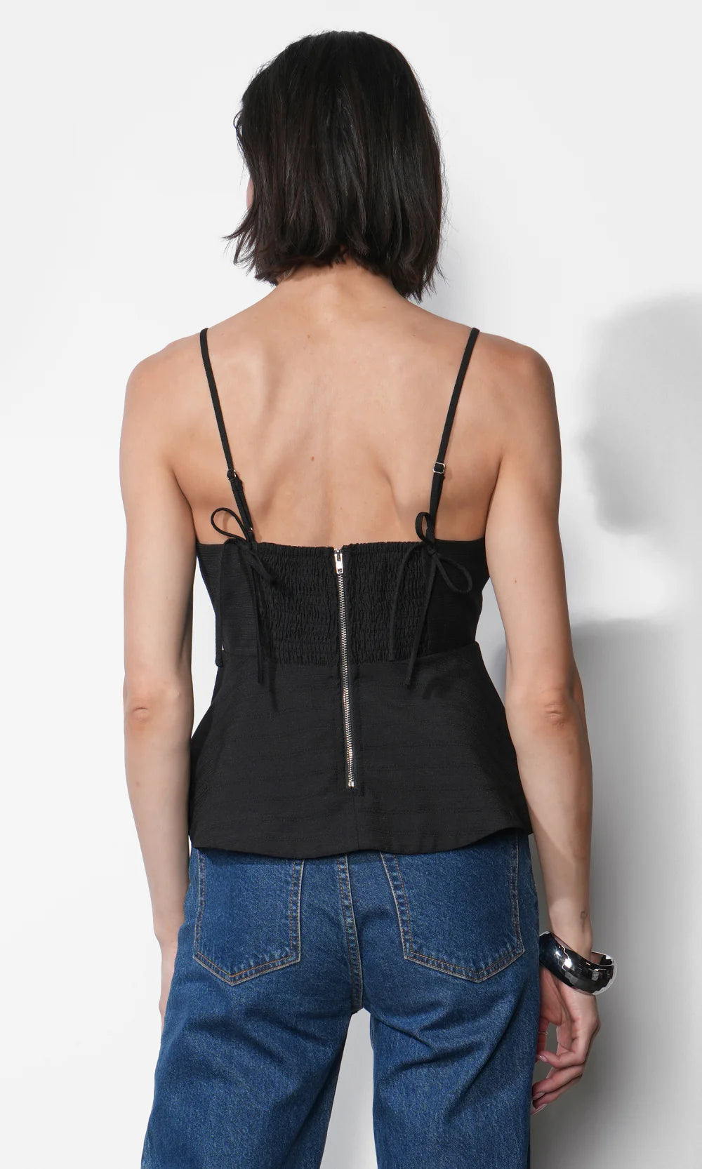 Person wearing a black top with thin straps and a zipper, paired with blue jeans on a white background. back view
