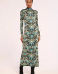A woman modeling a long-sleeve, high-neck maxi dress with an intricate paisley pattern in black, teal, blue, and beige, paired with black ankle boots against a solid light background.