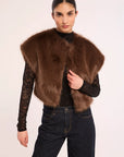 A model wearing a brown faux fur vest over a black lace long-sleeve top and dark blue jeans, photographed against a plain light background.