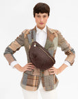 A model wearing a brown woven crossbody bag with gold hardware over a multicolor patchwork blazer and white shirt, against a plain white background.