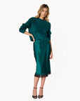 A woman modeling a dark teal green two-piece set consisting of a textured long-sleeve top and a satin midi skirt with a black lace hem, paired with black strappy heels against a plain white background.