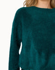 Close-up of a model wearing a fuzzy, long-sleeve knit sweater in a deep teal green color, paired with a matching satin skirt against a plain white background.