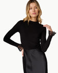 A woman modeling a black long-sleeve mock-neck top with sheer lace cuffs, paired with a black satin skirt, against a plain white studio background.