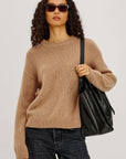 Woman wearing a beige sweater and dark jeans, holding a black bag against a plain background