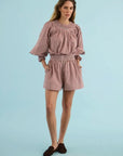 A woman modeling a pink and white gingham two-piece set consisting of a long-sleeve blouse with ruffled sleeves and matching shorts, paired with dark brown suede loafers, against a solid light blue studio background.