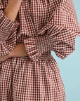 A close-up of a person wearing a red and white gingham dress with voluminous ruffled sleeves and a smocked waist, set against a solid light blue background.