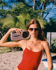 Woman in Heidi 12K Matte Black Fade Chroma + Blonde Venezia Sunglasses and a red swimsuit holding a vintage radio on a beach with palm trees in the background