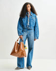 Person wearing a denim jacket and jeans holding a brown leather bag on a white background