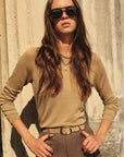 Woman wearing sunglasses and a beige sweater against a stone wall.